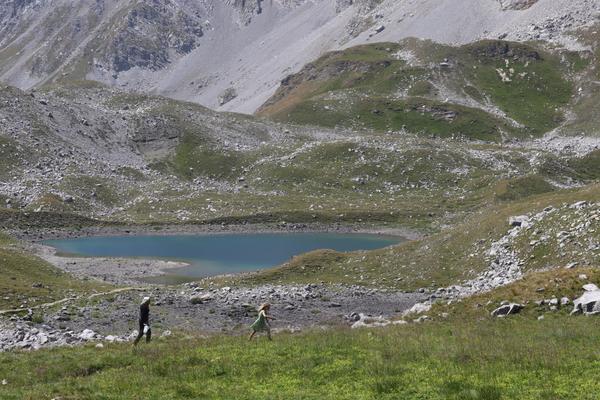 Two individuals are seen walking across a grassy, rocky landscape towards a serene blue mountain lake